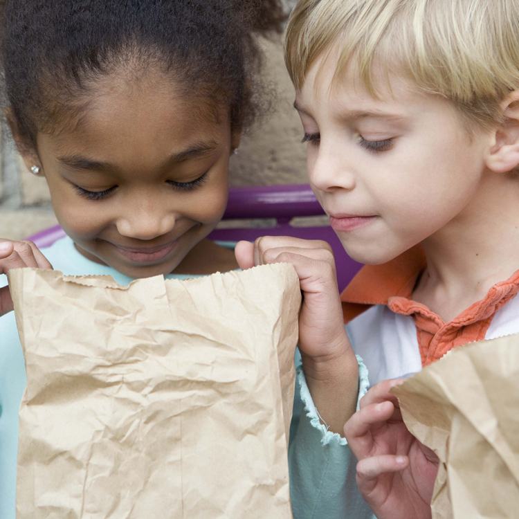 Two children looking into a brown paper lunch bag.